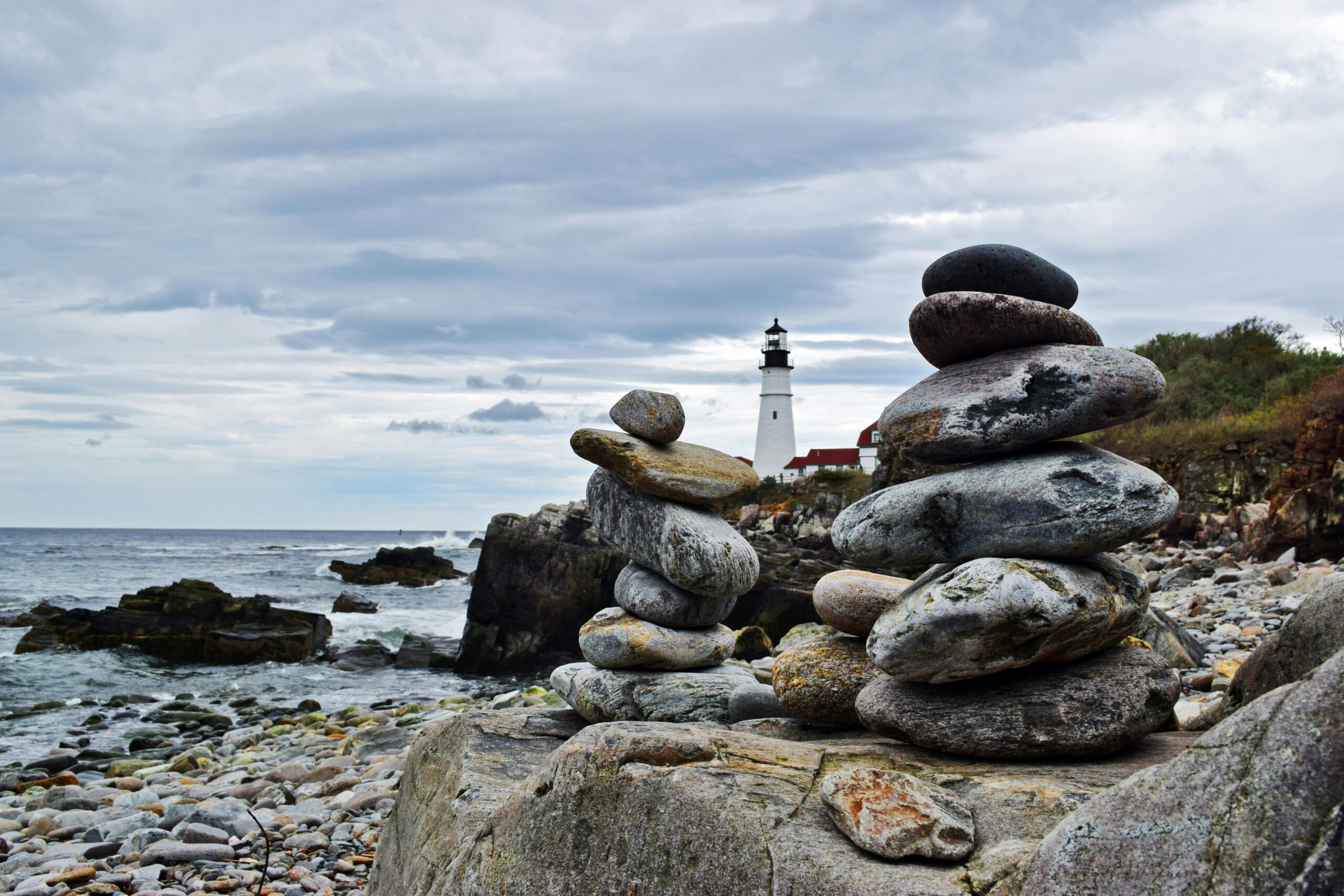 Stacked rocks by the coast with a lighthouse in the background, serene seascape.